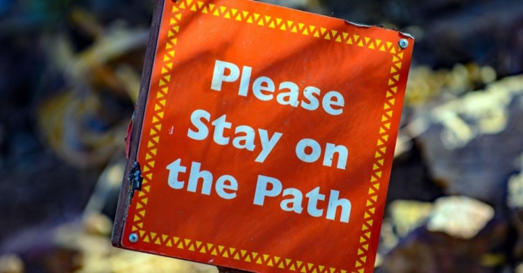An orange sign that says in white letters Please Stay On The Path