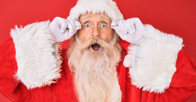 An old man dressed as Santa Claus and gripping his spectacles with a shocked expression
